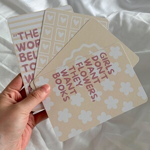 Könnte beinhalten: Ein Set aus drei rechteckigen Karten in einer cremefarbenen und beigen Farbpalette. Eine Karte zeigt den Text "GIRLS DON'T WANT FLOWERS, THEY WANT BOOKS" mit weißen Blumen-Icons. Eine weitere Karte hat ein Herzgitter und die dritte horizontale Streifen.
