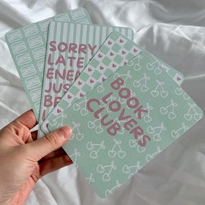 Peut inclure: Un ensemble de cartes rectangulaires aux coins arrondis avec des motifs vert pastel et blanc. Une carte affiche le texte BOOK LOVERS CLUB en rose avec un motif de cerises, tandis qu'une autre montre SORRY LATE ENERGY JUST BEING ME en rose.