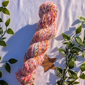 Peut inclure: Une écheveau de fil filé à la main aux couleurs rose, orange, jaune et vert. Le fil a un aspect texturé et est étiqueté avec une étiquette marron qui dit "sew fae handspun yarn".