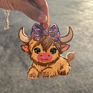 May include: A brown and white highland cow ornament with a purple glitter bow. The ornament is made of resin and has a textured surface.