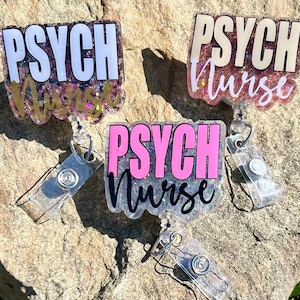 Puede incluir: Tres portagajes de brillo con carretes retráctiles. Cada portagajes presenta el texto "PSYCH NURSE" en diferentes colores y fuentes. Los portagajes están sobre una superficie gris texturizada.