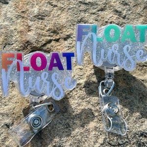 Peut inclure: Deux porte-badges transparents avec les mots "FLOAT Nurse" en lettres colorées et scintillantes. Le mot "FLOAT" est en orange, violet et bleu. Le mot "Nurse" est en écriture blanche. Les bobines ont un clip en plastique transparent et un cordon rétractable.
