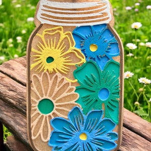 May include: A wooden mason jar-shaped decoration with a black metal loop for hanging. The jar is filled with colorful, layered flower cutouts in yellow, blue, and green. The background is a blurred view of a wooden surface and greenery.