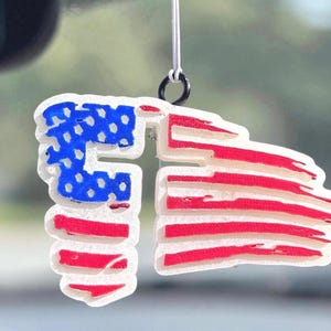May include: A patriotic car air freshener shaped like the American flag. The design features a white outline with red and white stripes and a blue field of stars. It hangs from a white string and black ring.