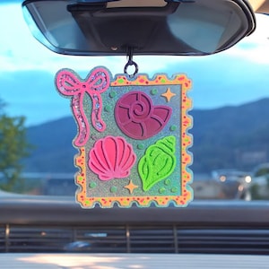 May include: A decorative car charm featuring a square design with a pink bow, a pink seashell, a purple seashell, and a green seashell. The charm has a glittery background and a yellow border. The charm is hanging from a car's rearview mirror.