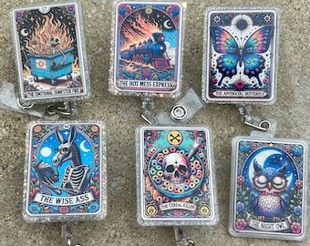 May include: Six glittery badge reels with tarot-inspired designs. The designs include a skeleton in a dumpster fire, a train, a butterfly, a donkey, a skull in cereal, and an owl. Each reel has a unique title, such as 'The Hot Mess Express'.