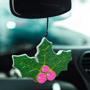 May include: A green glittery holly leaf car air freshener with pink berries, hanging from a black string. The air freshener is shaped like a holly leaf with white trim and is hanging from a car's rearview mirror.