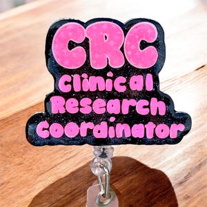 May include: A black badge reel with the text "CRC Clinical Research Coordinator" in pink. The letters "CRC" are in a bubble font with a star pattern. The badge reel has a clear plastic clip and a retractable cord.