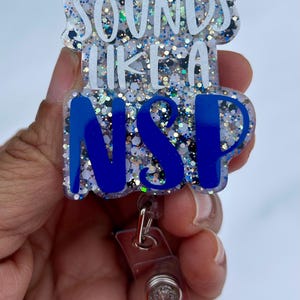 Könnte beinhalten: Ein transparenter glitzernder Badgehalter in Blau und Weiß mit einem silbernen, einziehbaren Clip. Der Badgehalter trägt den Text "Sounds like a NSP" in weißen Buchstaben.
