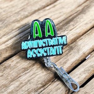 May include: A black and green badge reel with the text "AA ADMINISTRATIVE ASSISTANT" in a teal font. The badge reel has a clear plastic clip and a retractable cord. The background is a wooden surface.