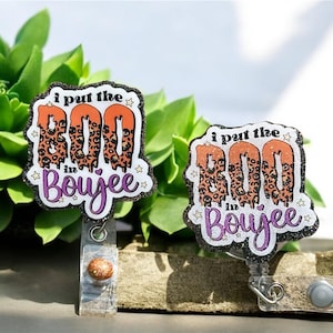 May include: Two badge reels with the text "I put the BOO in Boujee." The word "BOO" is orange with a leopard print pattern. The word "Boujee" is purple. The badge reels have a black glitter border and a clear plastic reel.