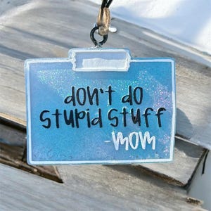 May include: A rectangular, light blue pendant with the text "don't do stupid stuff - mom" in black and white. The pendant has a glittery, sparkly finish and a white border. It is suspended by a black cord with a natural-colored twine accent.