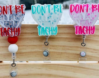 May include: Three heart-shaped badge holders with the text "Don't Be Tachy" in different colors. The badge holders are on a wooden surface.