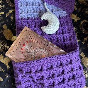 May include: A handmade crochet tarot card holder in shades of purple, lavender, and white. The holder features a pocket for cards and a decorative white crescent moon charm. A tarot card deck with celestial imagery is visible inside the pocket.