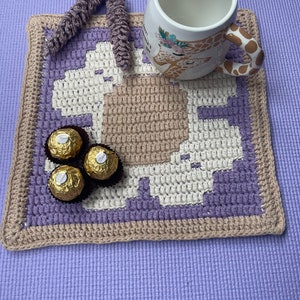 May include: A handmade crochet coaster in shades of purple, beige, and white, with a geometric pattern. A white ceramic mug with a giraffe design and three gold-wrapped chocolates sit on the coaster. A crocheted loop hangs from the coaster.