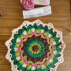 May include: A crocheted mandala featuring a brown, green and pink colour scheme. The mandala is crafted with a variety of stitches and has a delicate lace border.
