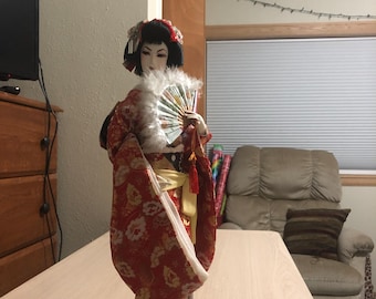 Pode incluir: Boneca gueixa japonesa em quimono floral vermelho, gola branca e leque. A boneca tem cabelo preto e está sobre uma base preta. É um item decorativo.