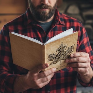 May include: A light brown wooden journal with a carved maple leaf design on the cover. The journal is open, revealing blank white pages. The text "Tree Sugar" is visible on the cover. The person holding the journal is wearing a red and black plaid shirt.