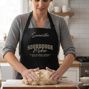 Pode incluir: Avental preto com o texto "Samantha" e "Sourdough Maker". O avental também tem o texto "Store bought bread? Bless your heart". A pessoa está amassando massa em uma tábua de madeira.