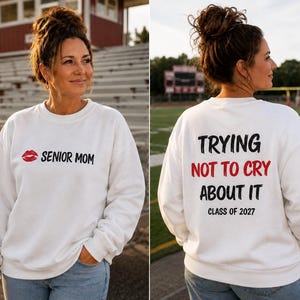 May include: A white long-sleeve sweatshirt featuring a red lip icon and the black text SENIOR MOM on the front. The back displays the black text TRYING NOT TO CRY ABOUT IT with the word NOT TO CRY in red, followed by CLASS OF 2027 in black.