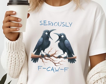 Pode incluir: Camiseta branca com um desenho de dois corvos pretos empoleirados num galho, com o texto azul "SERIOUSLY F-CAW-F". Os corvos têm o bico aberto. A camiseta é feita de um material macio.