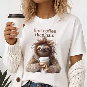 May include: White t-shirt with the text "first coffee then hair." The shirt features a cartoon sloth holding a coffee cup. The sloth has brown fur and a smiling face. The person is holding a white coffee cup.