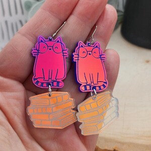 May include: A pair of dangle earrings featuring a bright pink cat wearing glasses on the top charm and a stack of orange books on the bottom charm. The cat and book charms are connected by silver-toned metal jump rings and attached to silver-toned earring hooks.