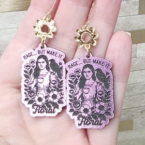May include: A pair of glittery pink rectangular earrings featuring a black illustration of a person holding a cup with a bird on their shoulder, surrounded by flowers. The text reads RAGE... BUT MAKE IT Floral. Gold-toned flower-shaped hooks.