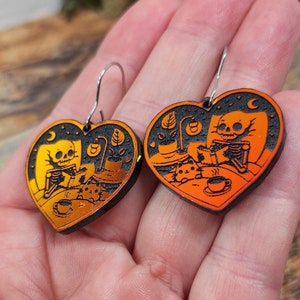 May include: A pair of heart-shaped dangle earrings featuring a vibrant orange and black engraved design. The illustration depicts a skeleton reading a book in bed with a cat, a lamp, and a coffee cup. The earrings have silver-toned metal hooks.