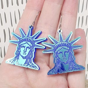 May include: A pair of blue Statue of Liberty earrings. Each earring features a detailed depiction of the iconic statue's face and crown, with a light blue and dark blue color scheme. The earrings hang from silver-colored hooks.