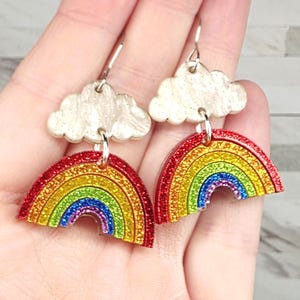 May include: A pair of dangle earrings featuring a rainbow design. Each earring has a red, orange, yellow, green, blue, and purple rainbow shape with glitter accents, connected to a silver-colored cloud charm.