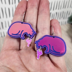 May include: A pair of pink and purple cat earrings with a dripping pink and purple design. The earrings are made of acrylic and have a shiny finish.