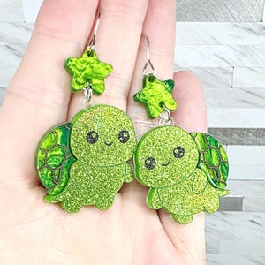 May include: A pair of glittery green turtle earrings with star-shaped accents. The earrings feature a smiling turtle design with a textured shell and are suspended from silver-colored hooks.