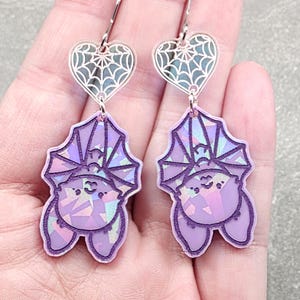 May include: A pair of purple bat earrings with silver heart-shaped tops. The bats have iridescent wings and smiling faces. The earrings are held in a hand against a gray background.