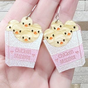 May include: A pair of novelty earrings featuring a white and pink box of chicken nuggets. Each earring has a box with the words "Chicken Nuggies" and a heart. The nuggets are yellow with orange beaks.