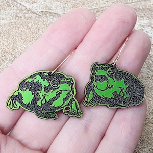 May include: A pair of frog-shaped earrings. Each earring features a black outline with a bright green frog design. The earrings are suspended from gold-colored ear wires.