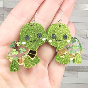 May include: A pair of green glitter turtle earrings with silver hooks. The turtles have black eyes, a black curved mouth, and a black and gold design on their necks. The turtle shells have a four-leaf clover design.