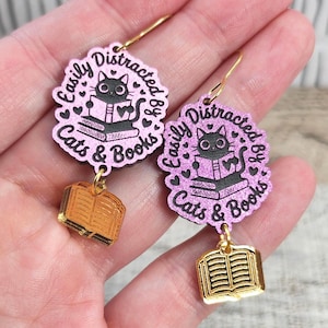 May include: A pair of dangle earrings featuring a pink glittery circular charm with a black cat reading a book illustration and the text Easily Distracted by Cats & Books. A small gold-toned metal book charm hangs from the bottom of each piece.