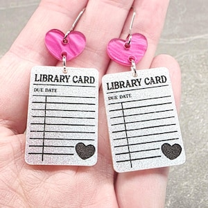 May include: A pair of dangle earrings featuring a rectangular silver glitter pendant printed with the text LIBRARY CARD and DUE DATE, along with black horizontal lines and a small heart. Each earring has a pink marbled heart-shaped top piece.