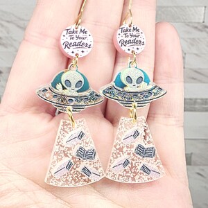 May include: Gold-tone dangle earrings featuring a pink and white circular charm with the text "Take Me To Your Readers". Below are blue and silver UFOs with alien figures, and a clear, glittery book-shaped charm.