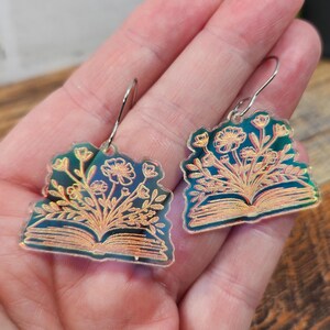 May include: A pair of dangle earrings featuring a dark teal, translucent acrylic base shaped like an open book. The surface is etched with a detailed copper-colored floral design emerging from the pages, suspended by silver-toned metal hooks.