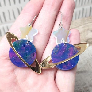 May include: A pair of space-themed earrings. Each earring features a blue and purple planet with a gold ring, a star-shaped top, and silver hooks. The planets have a galaxy-like design with glitter.