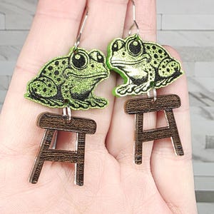 May include: A pair of whimsical earrings featuring green frog designs with black spots. Each frog is perched above a small, brown wooden stool. The earrings have silver-colored hooks.