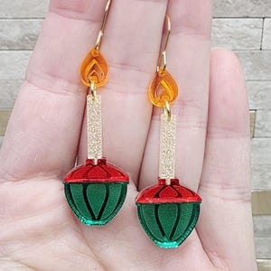 May include: A pair of festive earrings featuring a red and green ornament design. The earrings have gold-colored hoops and accents, with a dangling orange flame-shaped top. The ornament portion is green with red accents.