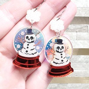 May include: A pair of novelty earrings featuring a snow globe design. Each earring has a clear, circular globe with a snowman wearing a top hat and a skull face. The base is red, and the top has a white, snowflake-shaped accent.