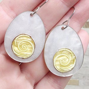 May include: A pair of silver-tone teardrop earrings with a gold-colored spiral design. The earrings have a pearlescent, light gray background and a simple hook closure. The gold spiral is centered on each earring.