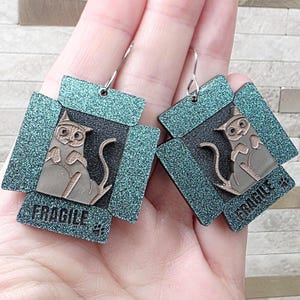 May include: A pair of cat-themed earrings. Each earring features a gray cat illustration inside a box with the word "FRAGILE" printed on the bottom. The box has a teal glittery border and silver-colored ear wires.