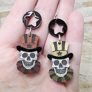 May include: A pair of Halloween-themed earrings. Each earring features a skull with a top hat and a star-shaped accent. The hats are decorated with glitter. The skull has a ruffled collar. The earrings are suspended from a black circle with a star cutout.