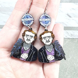 May include: A pair of dangle earrings featuring a portrait of Edgar Allan Poe with a raven perched on his shoulder. Above the portrait is a circular charm with a purple galaxy pattern and the word NEVERMORE printed in white capital letters.