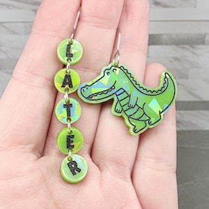 May include: A pair of novelty earrings. One earring spells out the word "LATER" in green circular beads. The other earring is a green alligator with a geometric design. Both earrings have silver-colored hooks.